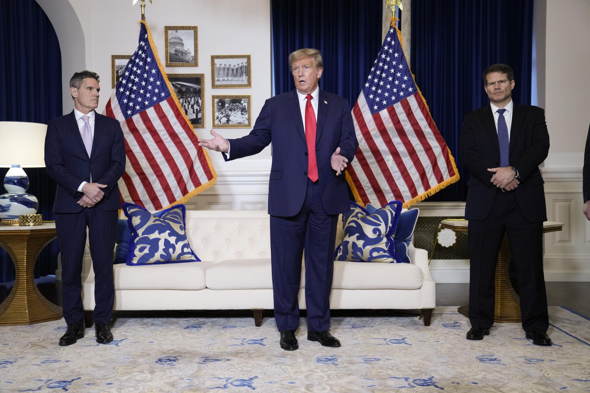 Former President Trump, flanked by two men in suits, stands and speaks in a white room with blue accents and two U.S. flags