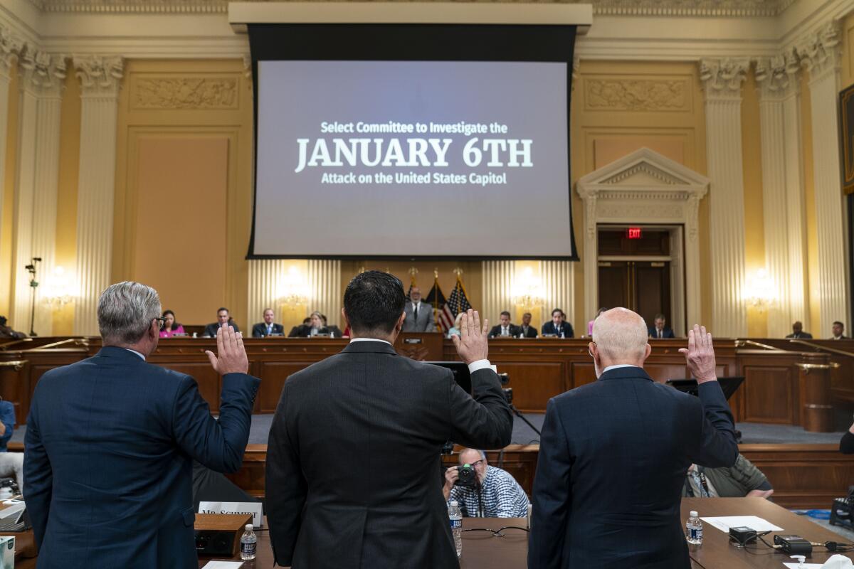 Three men in dark suits with their backs to the camera raise their hands to be sworn in to testify before a committee.