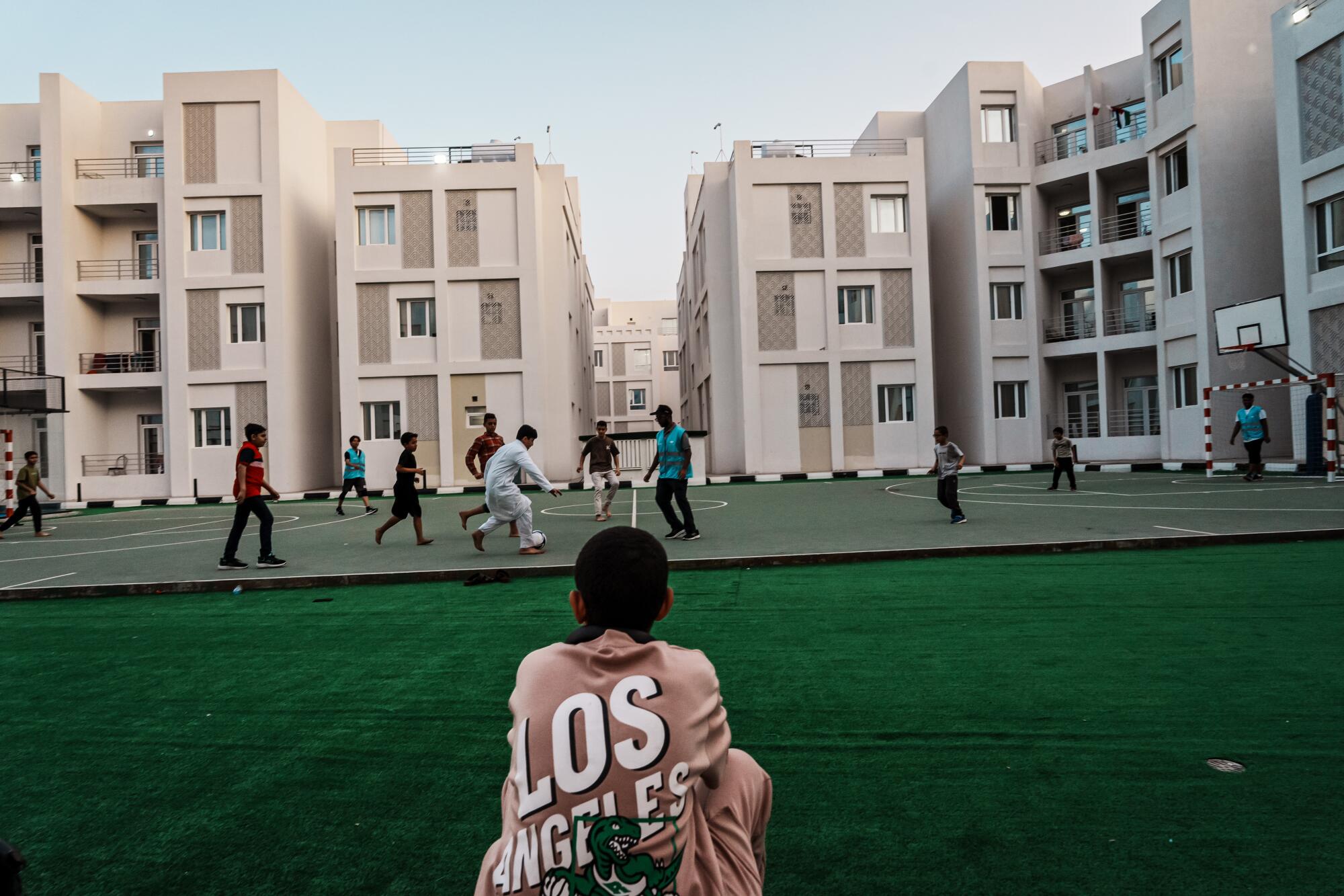 A boy wearing a T-Shirt that reads "Los Angeles" watches people playing soccer near buildings