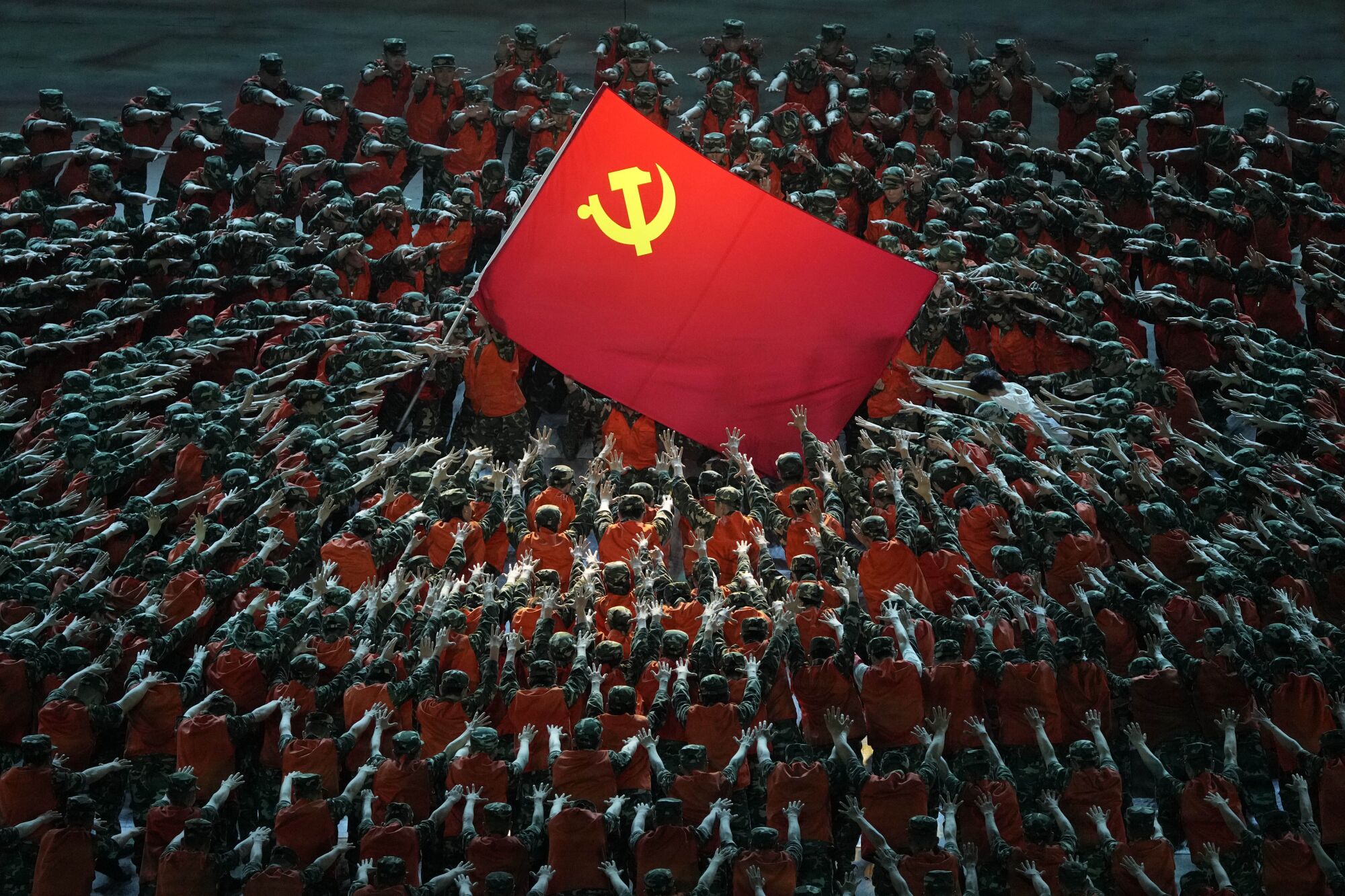 A large red flag with a hammer and sickle symbol is encircled by rows of people wearing red, with arms extended