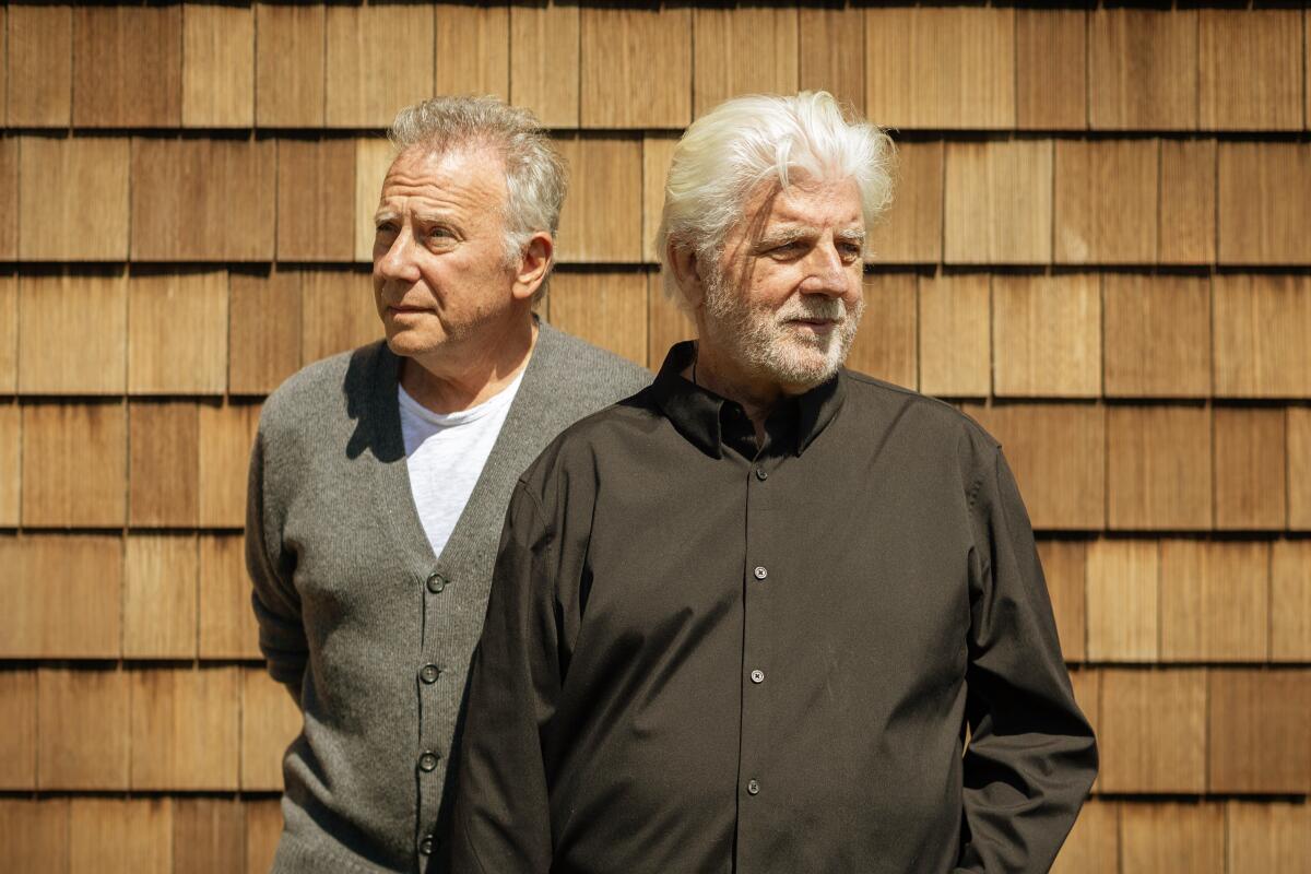 Two gray-haired men stand in front of a shingled wall