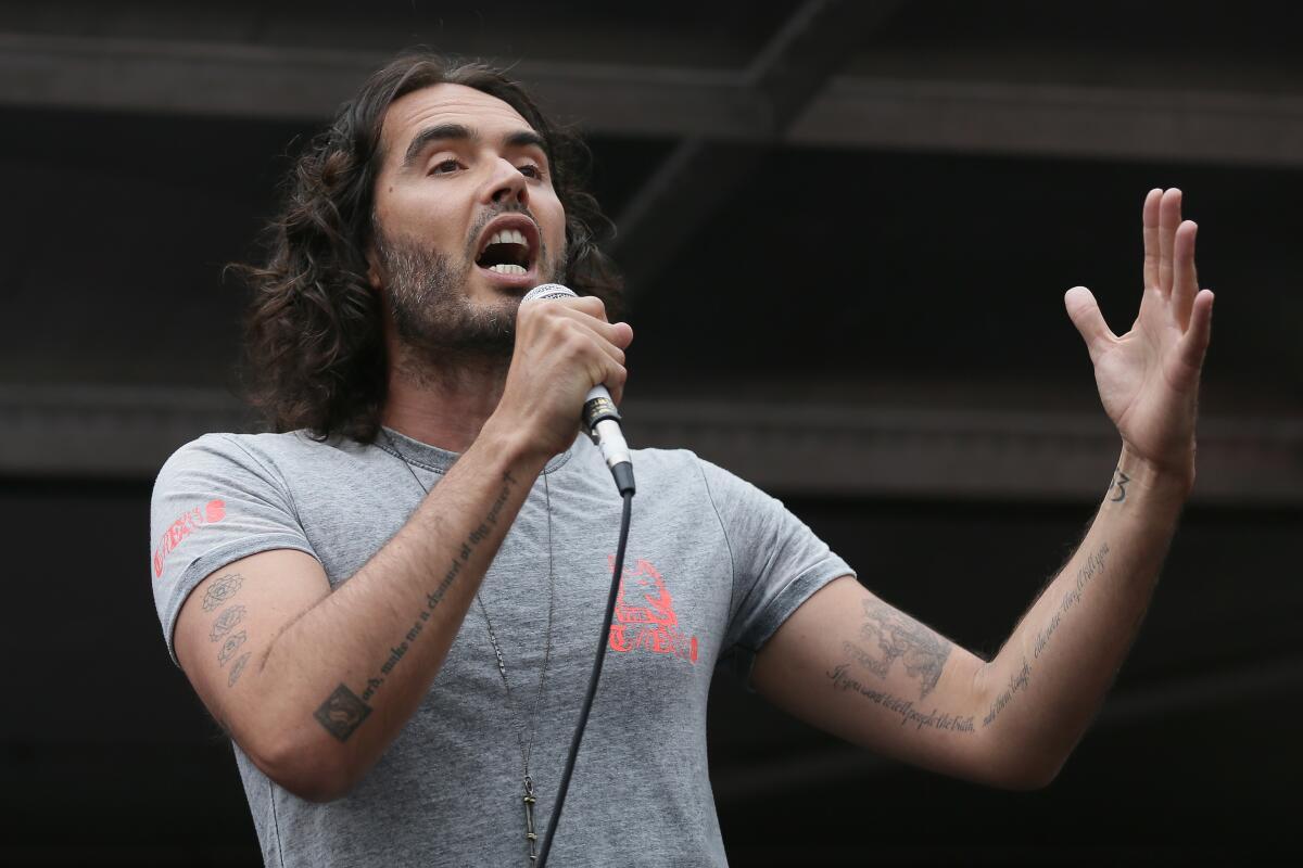Russell Brand wears a gray shirt as he speaks into a microphone while addressing a crowd.