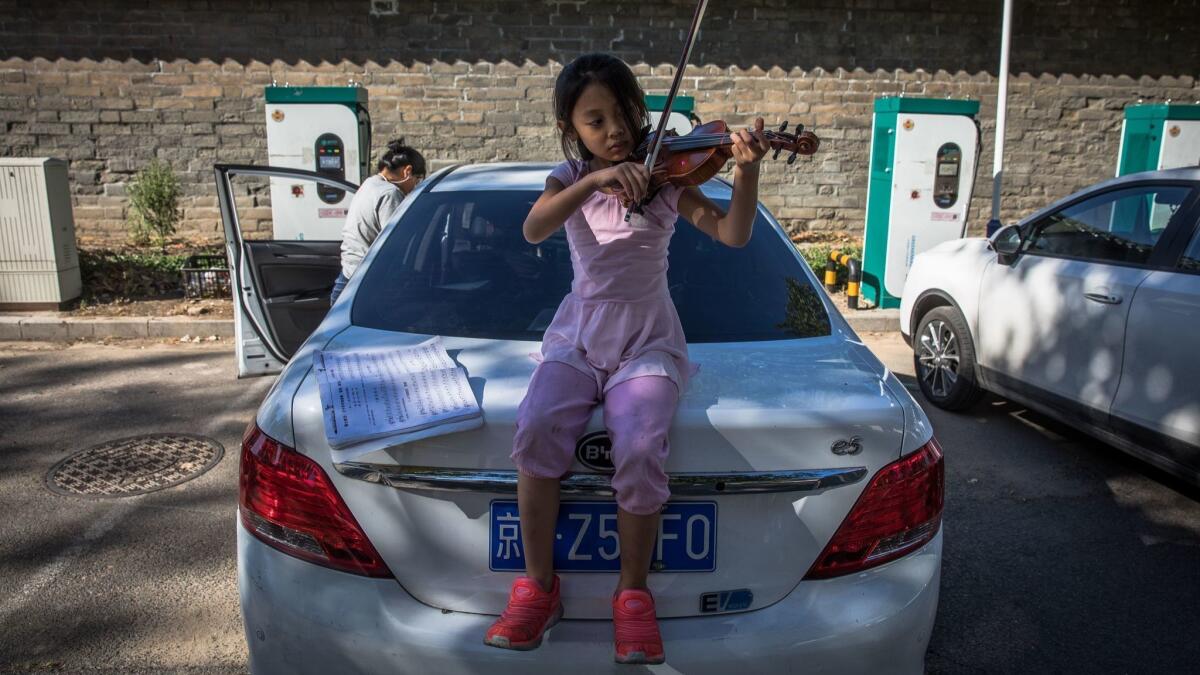 A Chinese girl plays the violin on a trunk of an electric car as her mother waits to charge it in Beijing.