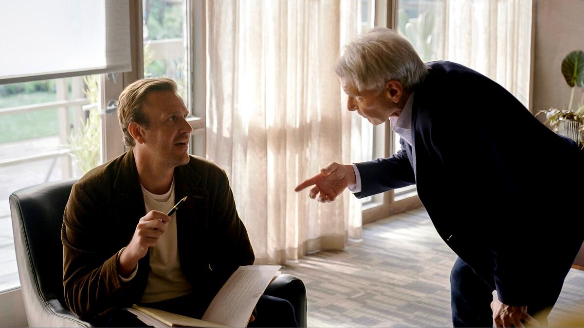 A man in a suit bends down to talk to a man in a chair with a notebook in his lap.