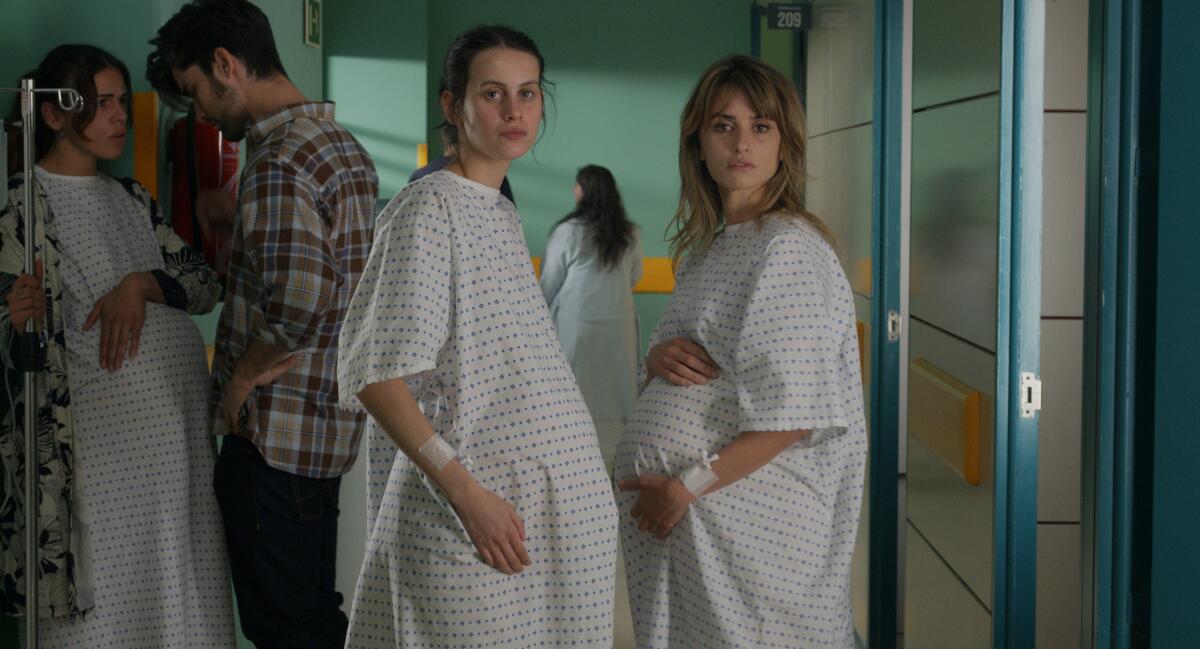 Two pregnant women stand in a hospital corridor, wearing smocks.