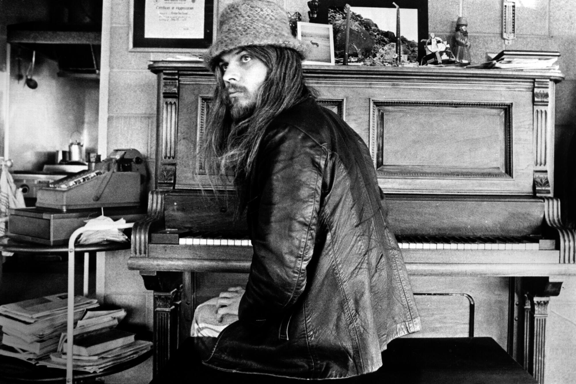 A singer-songwriter with long hair and a hat sits in front of a piano