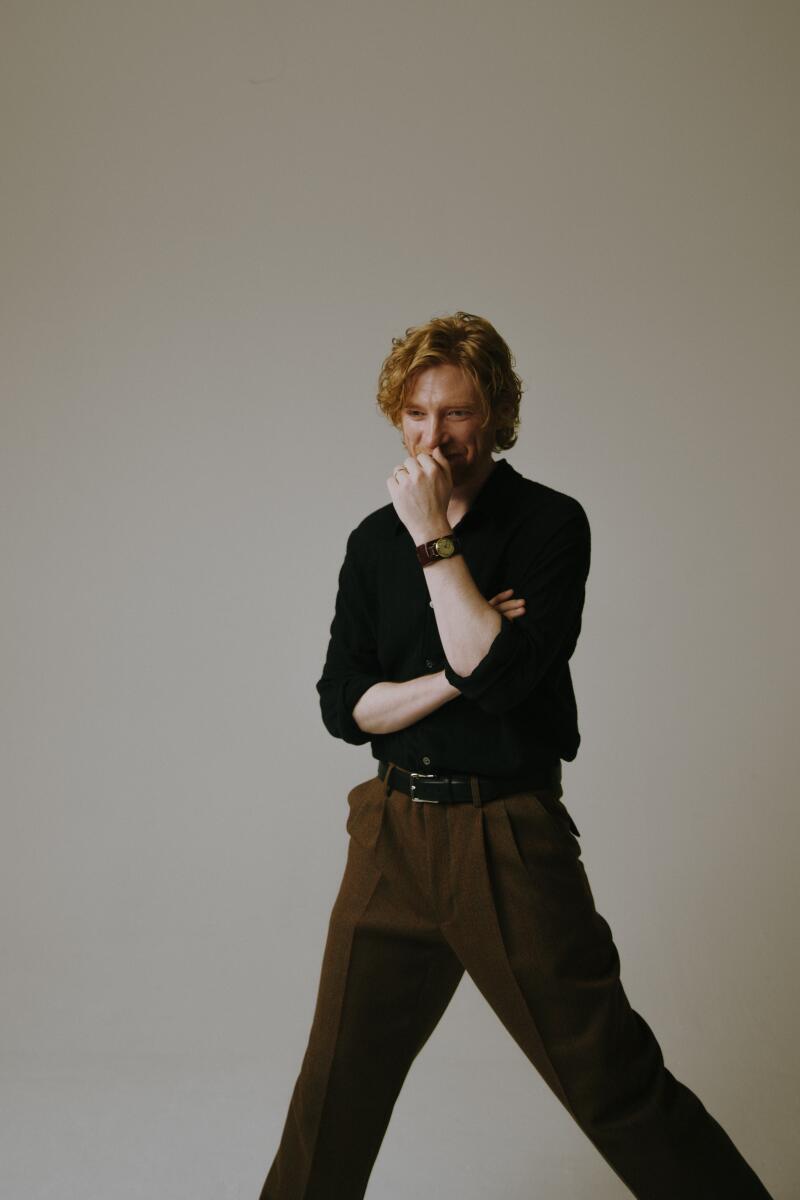A man stands with his legs wide and his left arm raised and clenched into a fist that covers his mouth.