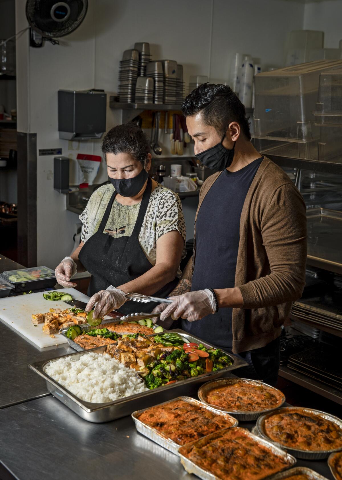 A mother and son, wearing masks, prepare large trays of food in a restaurant kitchen