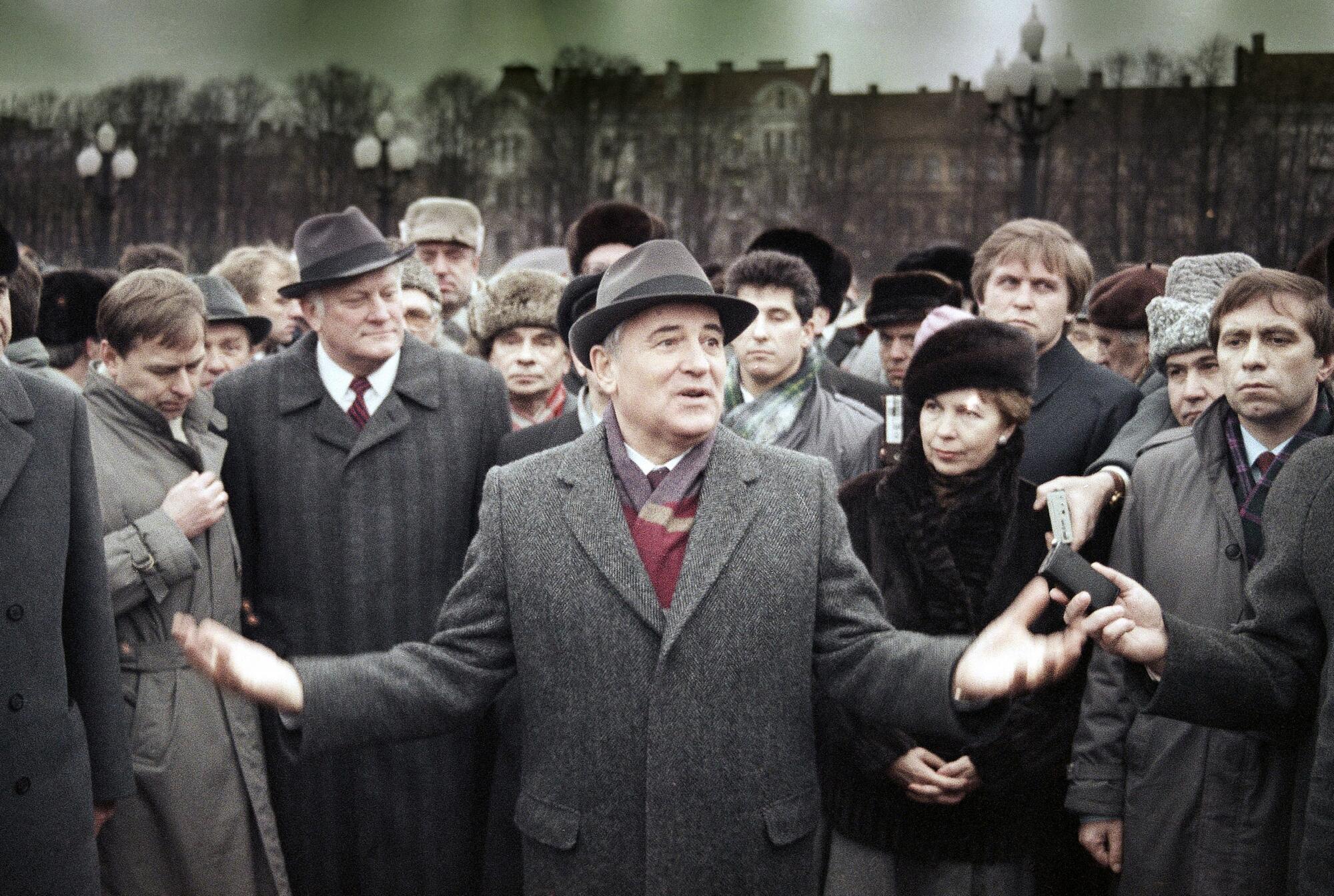 A man in a gray overcoat and hat, his hands apart with palms up, speaks to a crowd surrounding him 