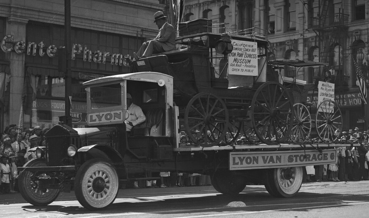 A boxy, 1930s-era truck carries on its flatbed two stagecoaches.