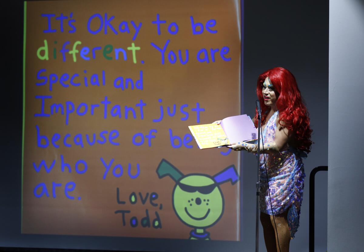 A man in drag speaks into a microphone while holding a book.