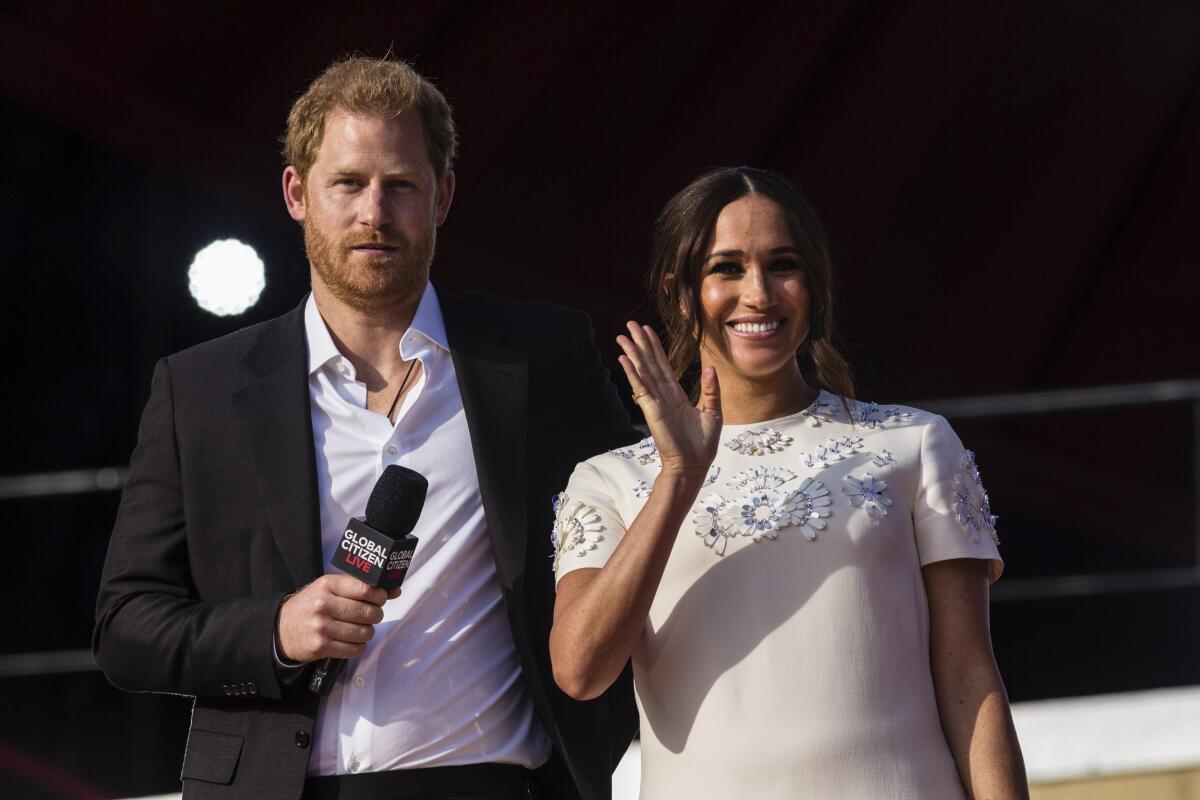 Prince Harry holds a microphone while standing next to Meghan, who smiles and waves.