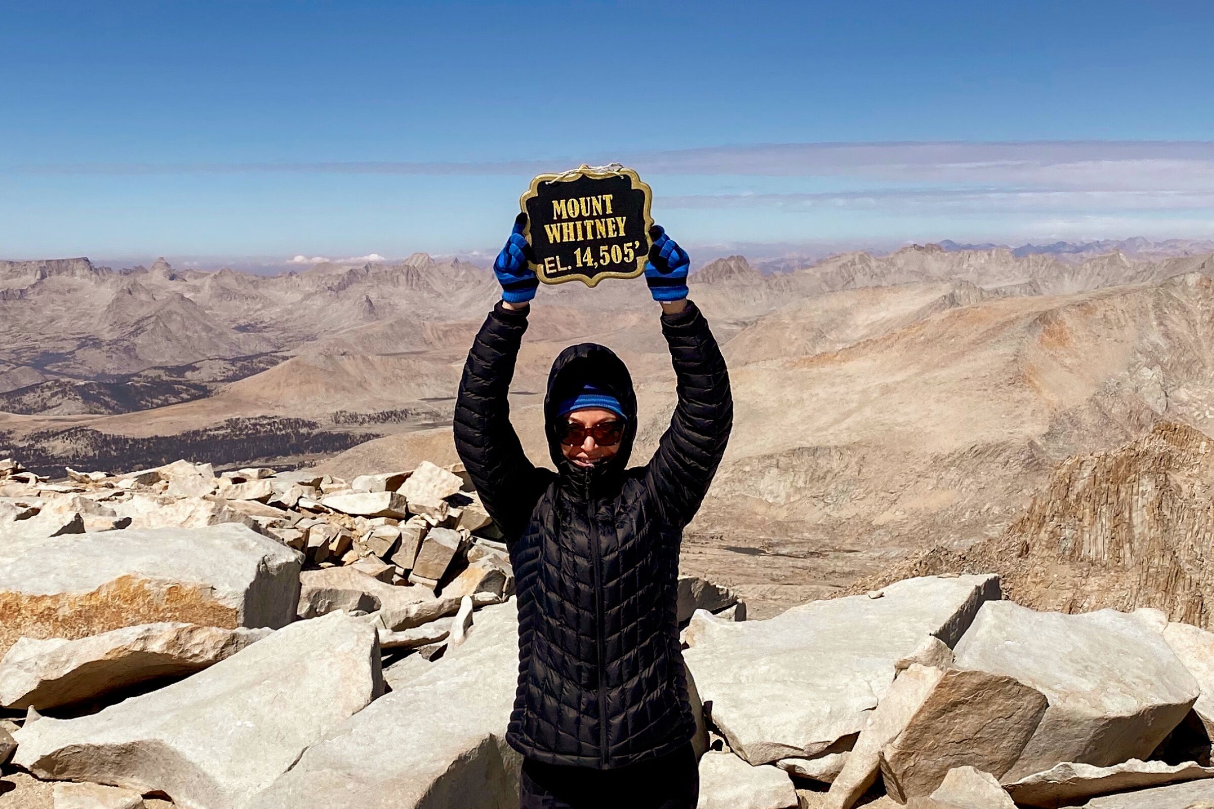 A woman on a mountaintop holding up a sign that says "Mount Whitney: Elevation 14,505 feet"