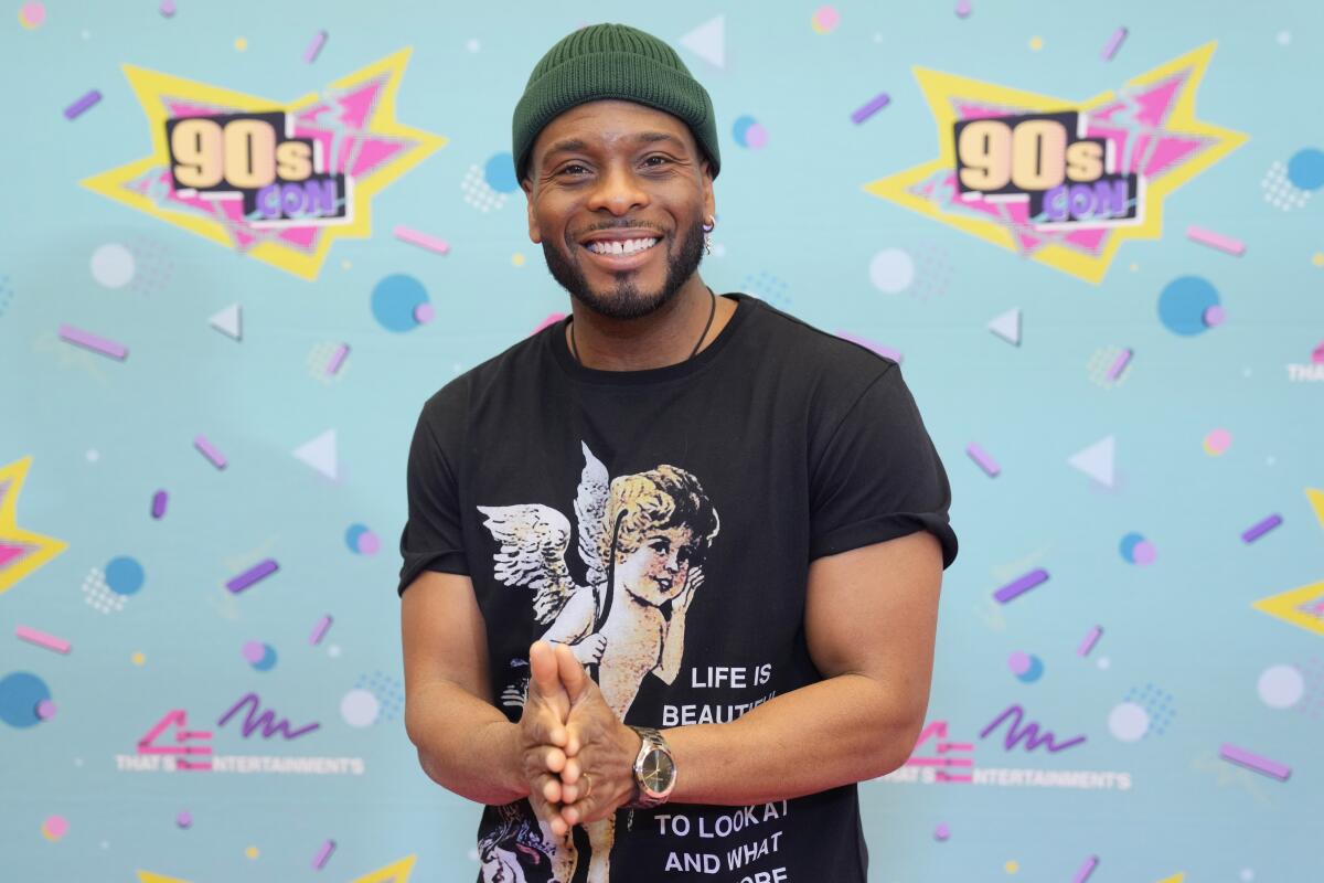 Kel Mitchell smiles with his hands together while posing in a green beanie and black graphic T-shirt with an angel