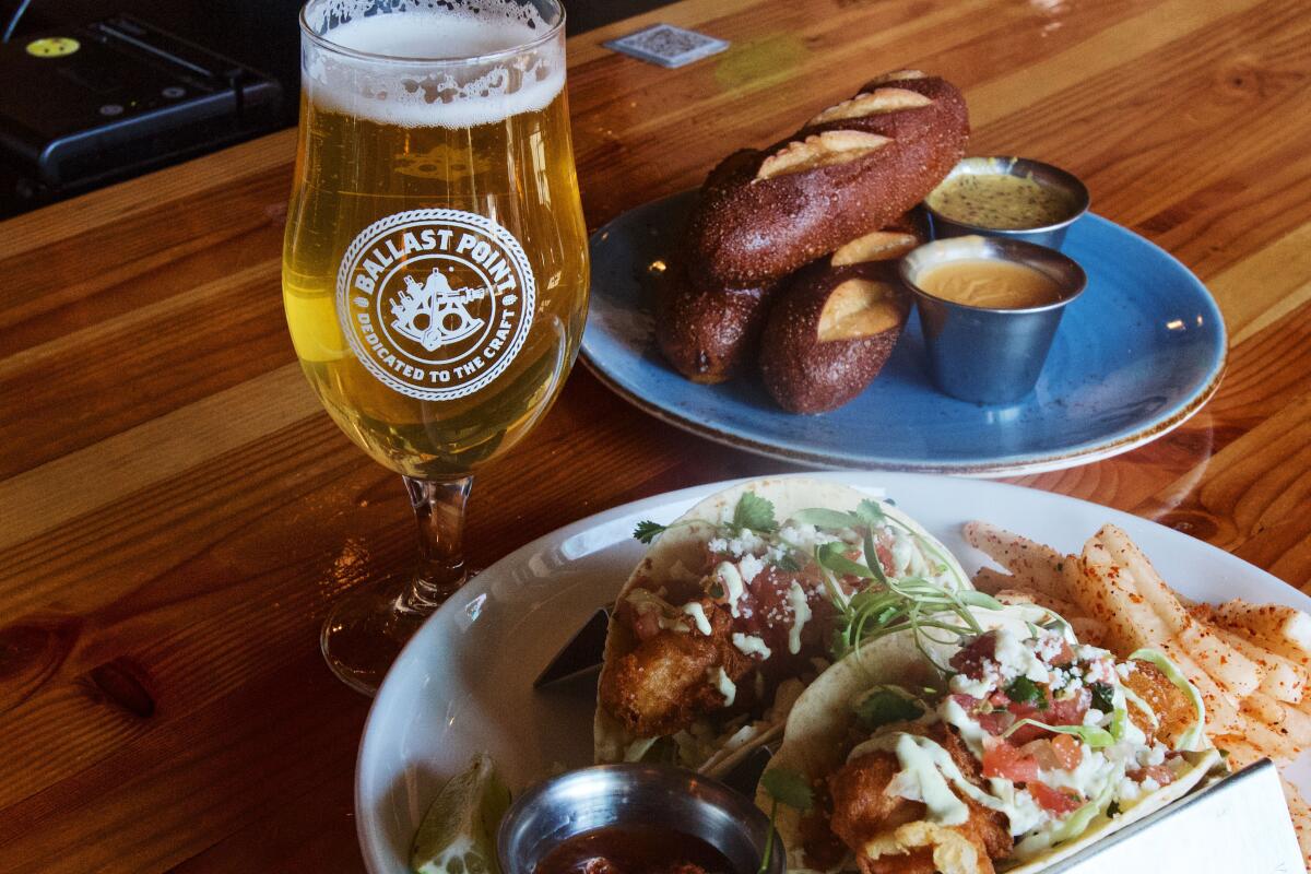 A tall golden beer to the left of two fried fish tacos and a stack of three pretzel loaves with beer cheese and beer mustard.