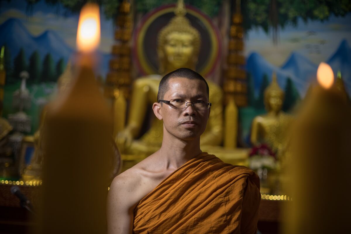 A Buddhist monk wearing glasses and orange robes looks into the camera with a stoic expression