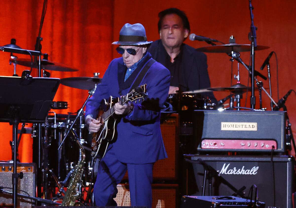 Van Morrison, in a blue suit and hat, plays guitar onstage