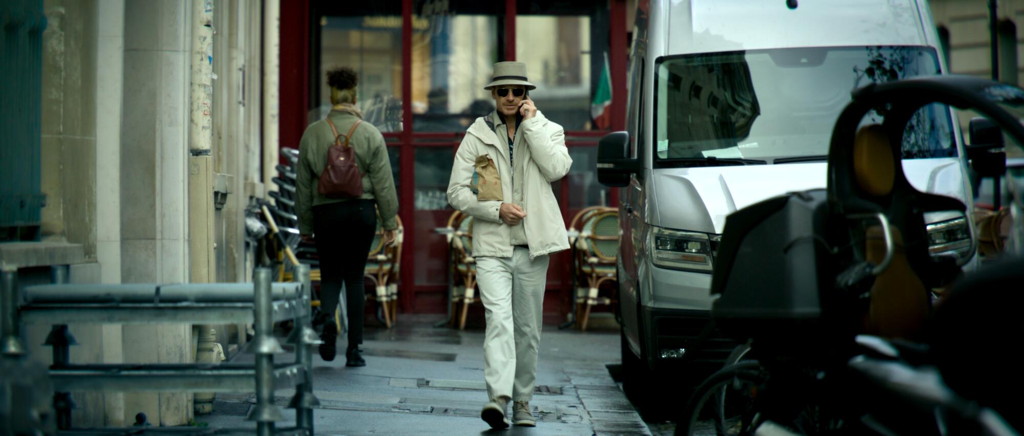 A man in shades and a white hat walks down a city street.