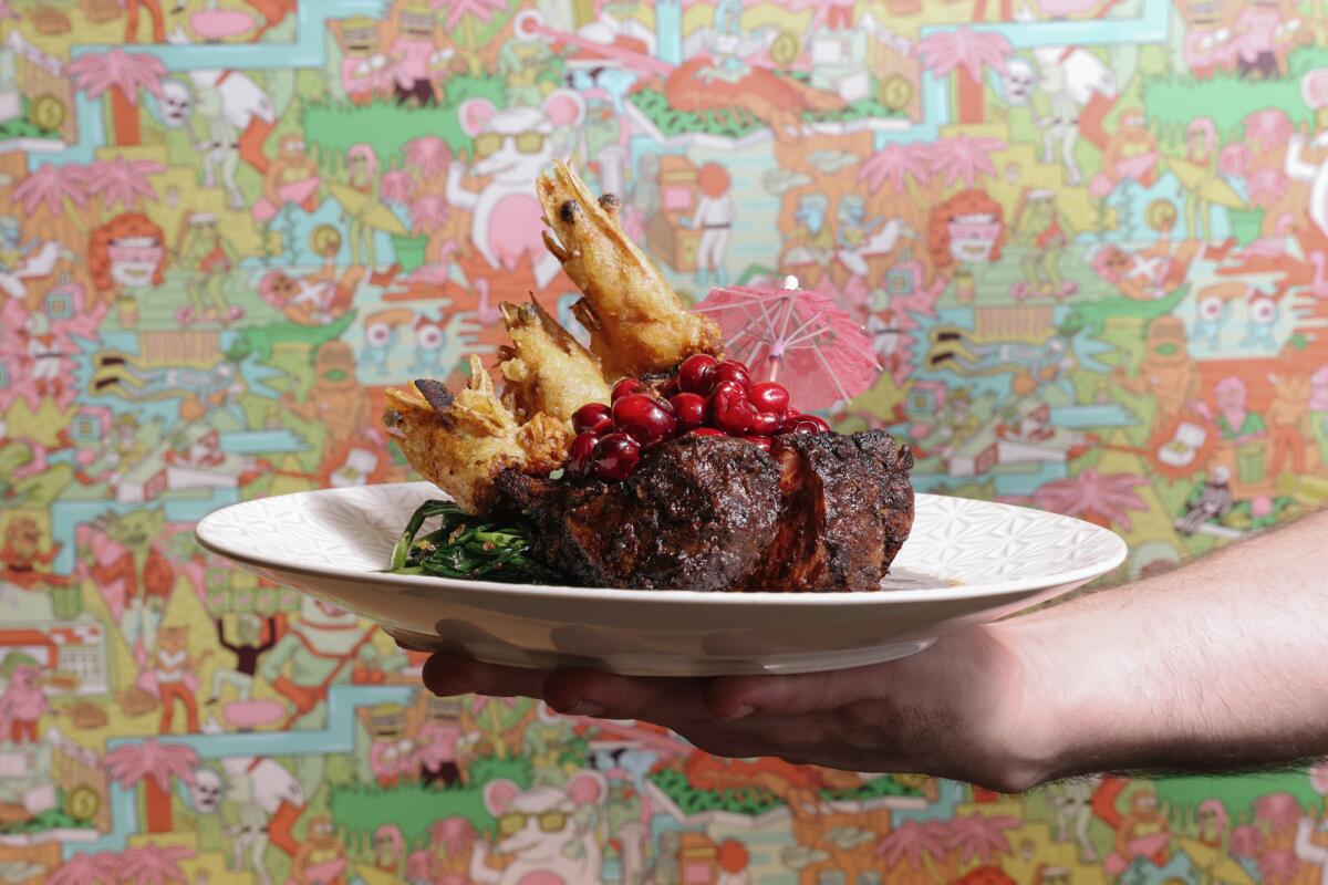 a hand holds out a plate with lamb chop and prawns in front of bright wallpaper 