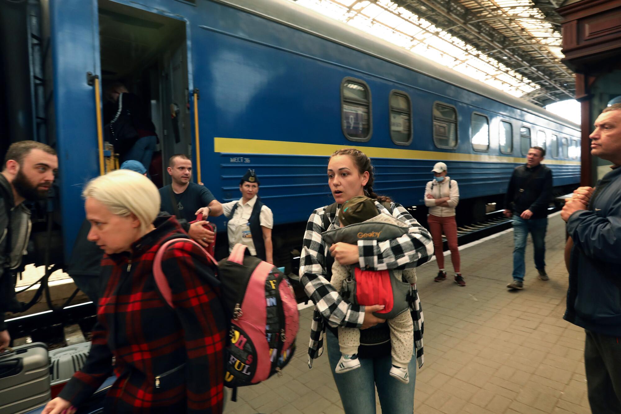 A woman with a backpack and another holding a baby are among the people disembarking from a blue train car with yellow stripe