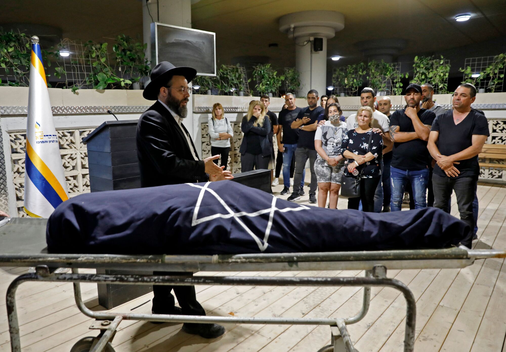 Mourners stand back from a wrapped body while a man stands next to it