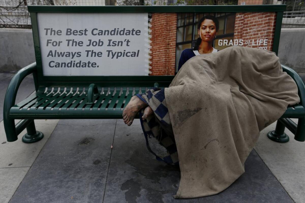 A homeless person sleeps under a blanket on a bus bench on South Grand Avenue.