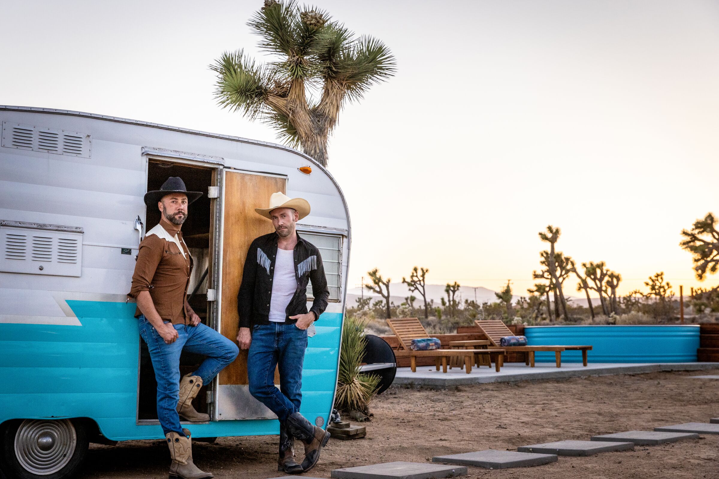 Two men in western garb and cowboy hats stand in front of a camper next to a stock tank pool.