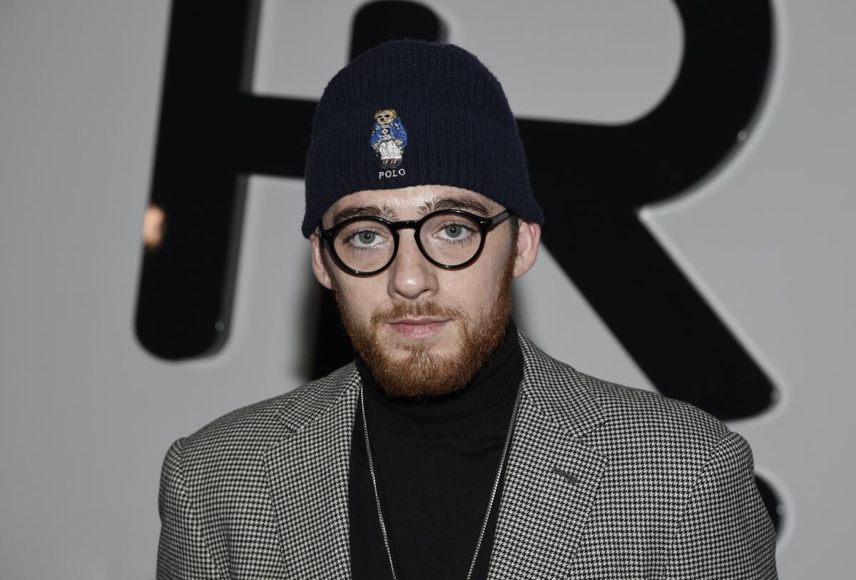 Angus Cloud in round glasses, a black beanie and turtleneck and a checker-print blazer