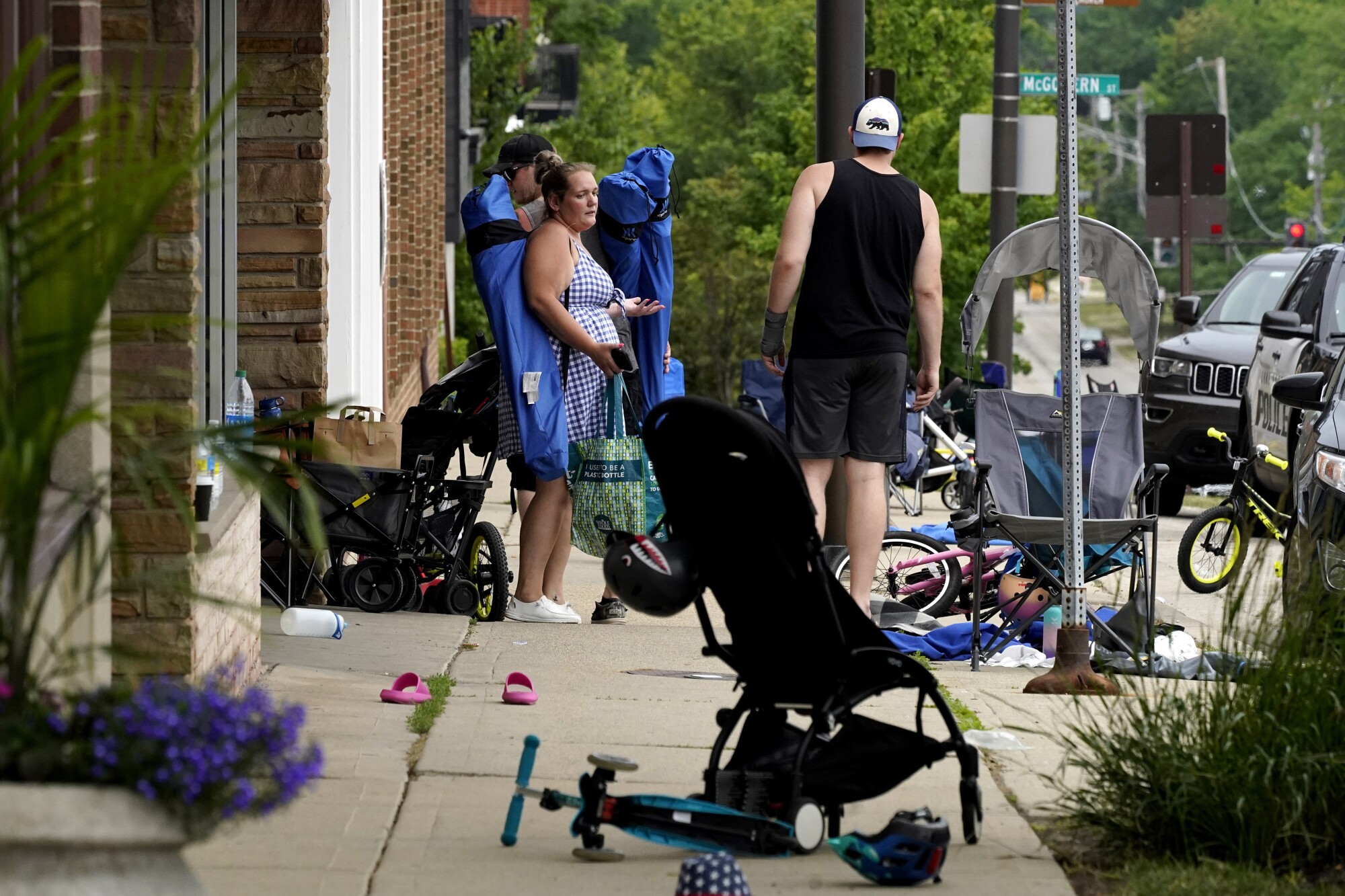 Several people stand on the sidewalk among a pile of empty chairs, strollers and scooters.
