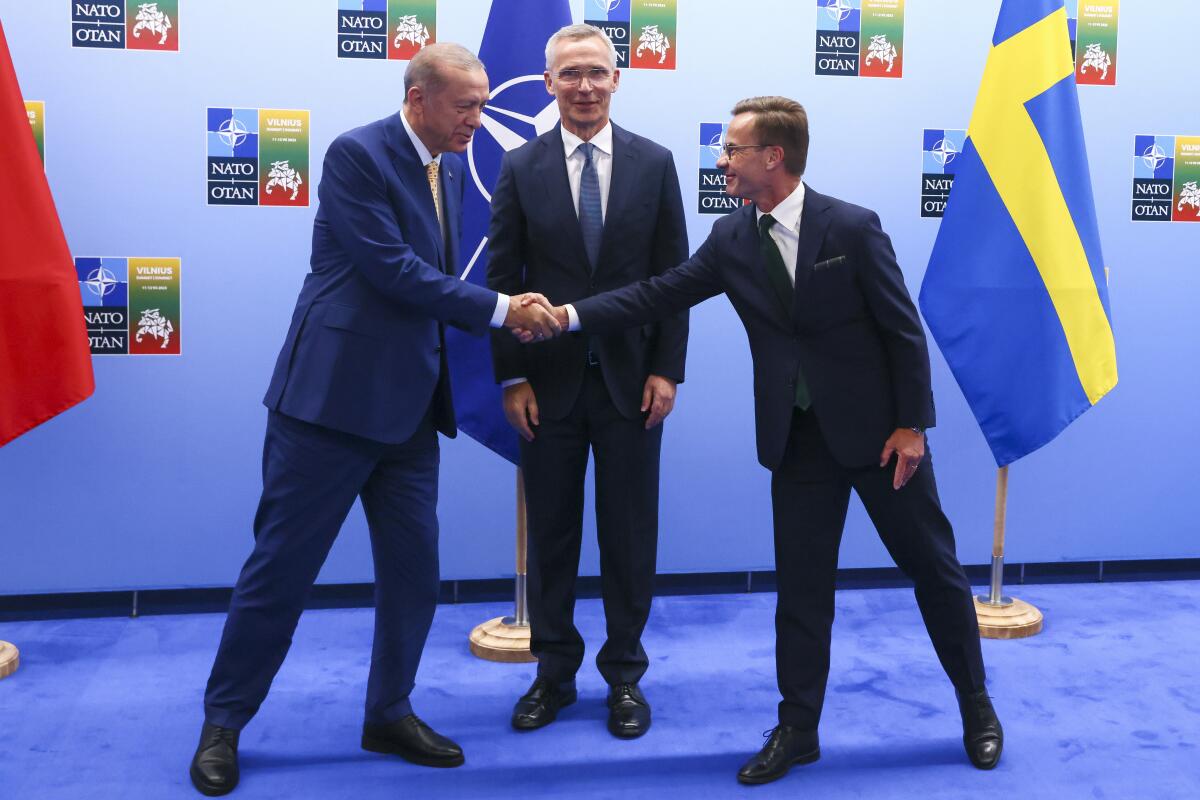 Two men in suits shake hands while another man looks on.