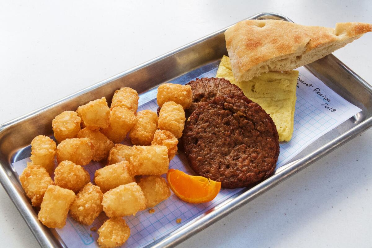 An angled photo of a vegan-egg omelet square, two round breakfast sausages, tater tots, a tiny citrus slice and focaccia