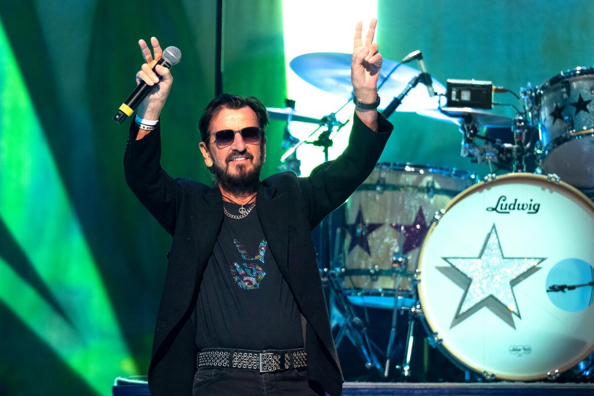 Ringo Starr wears sunglasses and flashes peace signs in front of a drum kit.