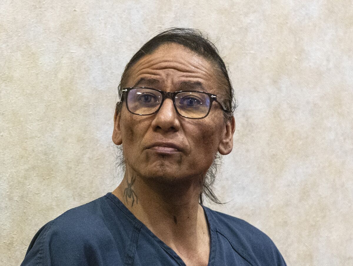 A man with a blue jailhouse shirt, glasses, marks on his chin and a tattoo on his neck sits in a courtroom