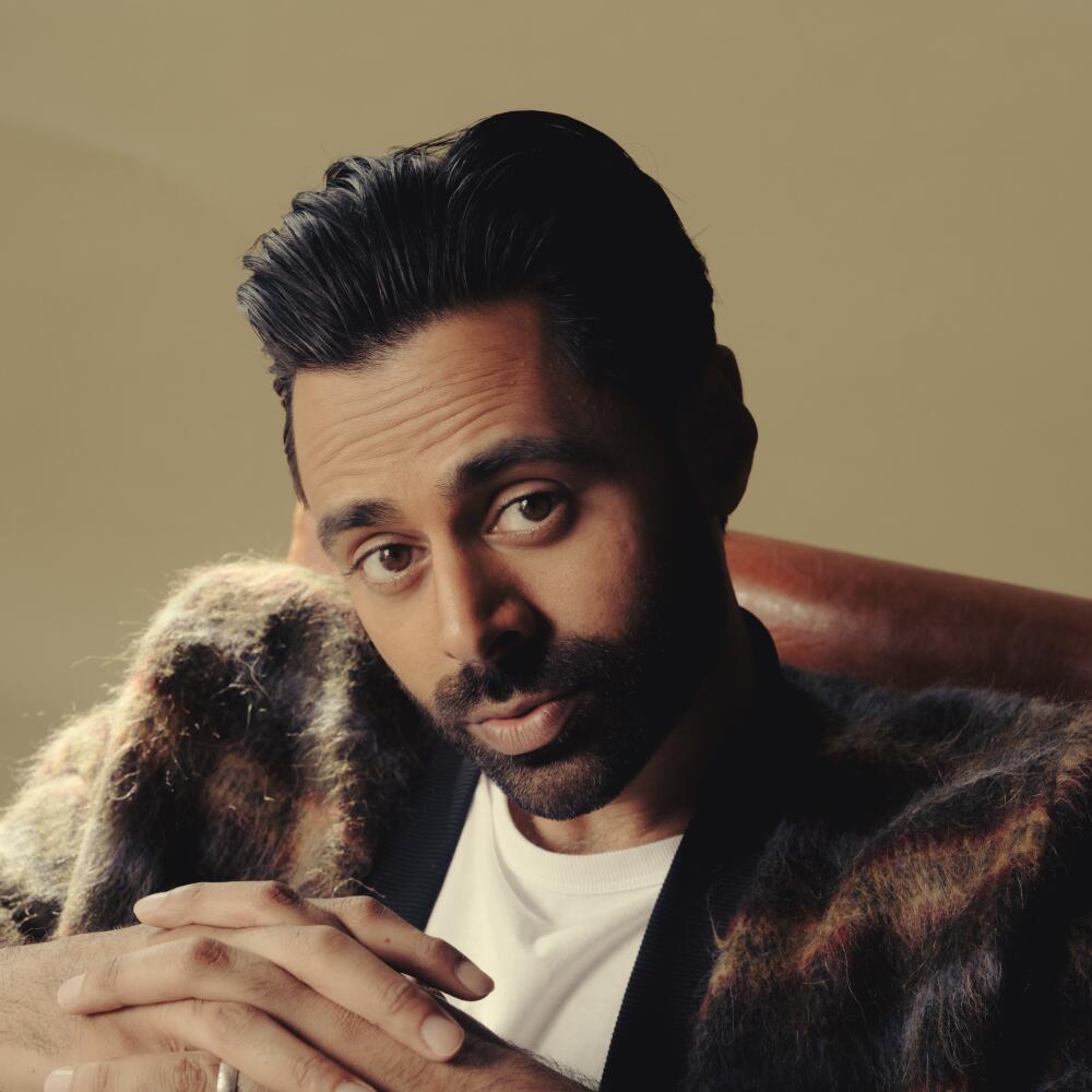 A man with dark hair in a white T-shirt and patterned sweater sits with his hands clasped.