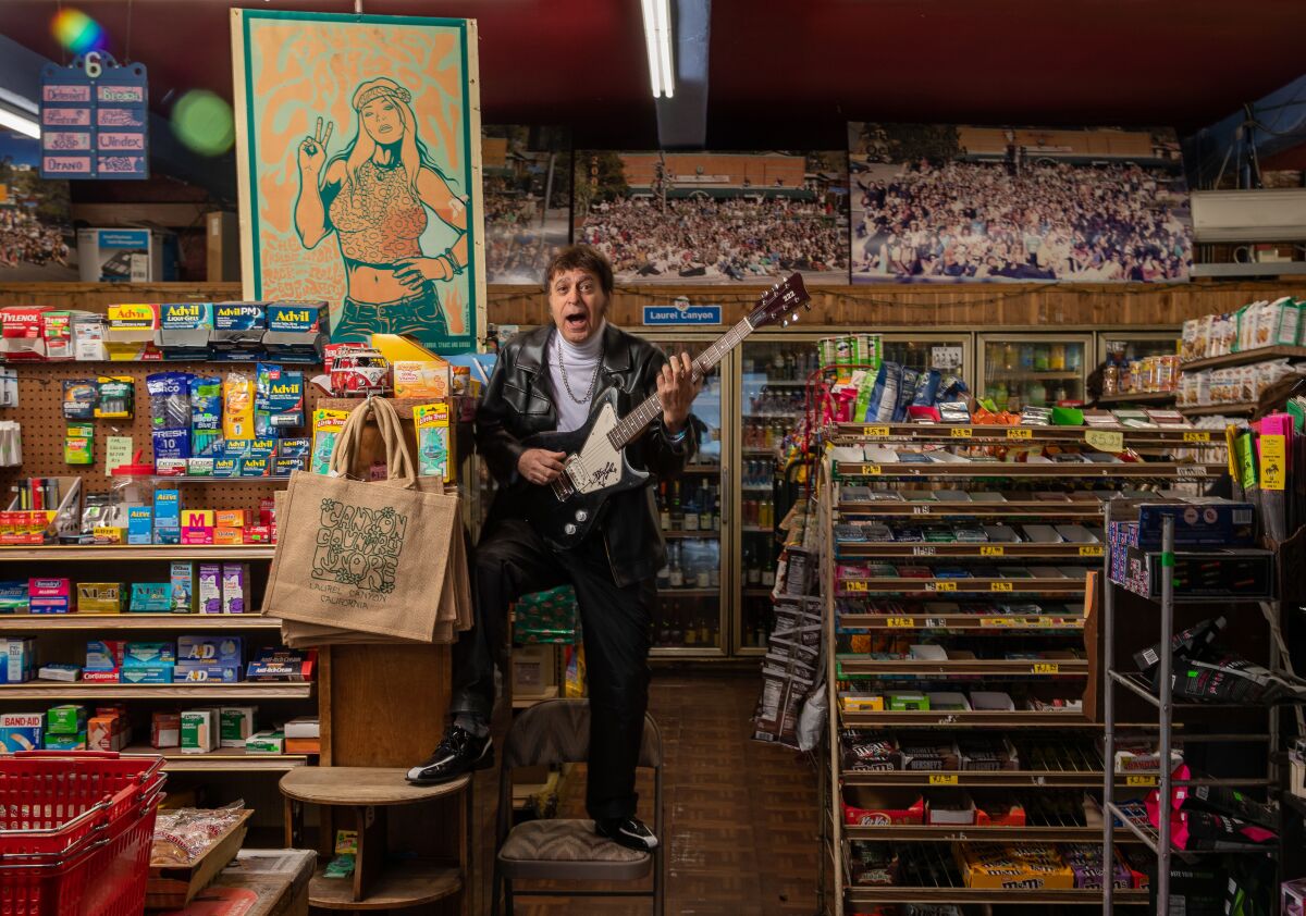 A man in a leather jacket stands and holds an electric guitar in a grocery aisle