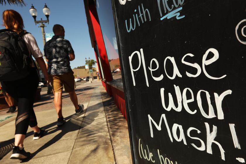 HUNTINGTON BEACH, CA - AUGUST 12, 2020 - - A couple walk past a sign that reads, "Please Wear Mask," in front of Dash of Sass clothing store along Main Street during a "Masks Up, Surf City," banner campaign to prevent coronavirus in the city of Huntington Beach on August 12, 2020. Customers are not allowed into the store unless they are wearing a mask. The campaign, a play on the phrase "Surf's Up," includes 100 pennants, a banner at the pier and digital displays citywide, Huntington Beach spokeswoman Catherine Jun said. Fifty pennants were installed downtown on Aug. 6. The remaining signs are in production this week, and 13 pennants and a banner are expected to be installed on the Huntington Beach Pier. (Genaro Molina / Los Angeles Times)