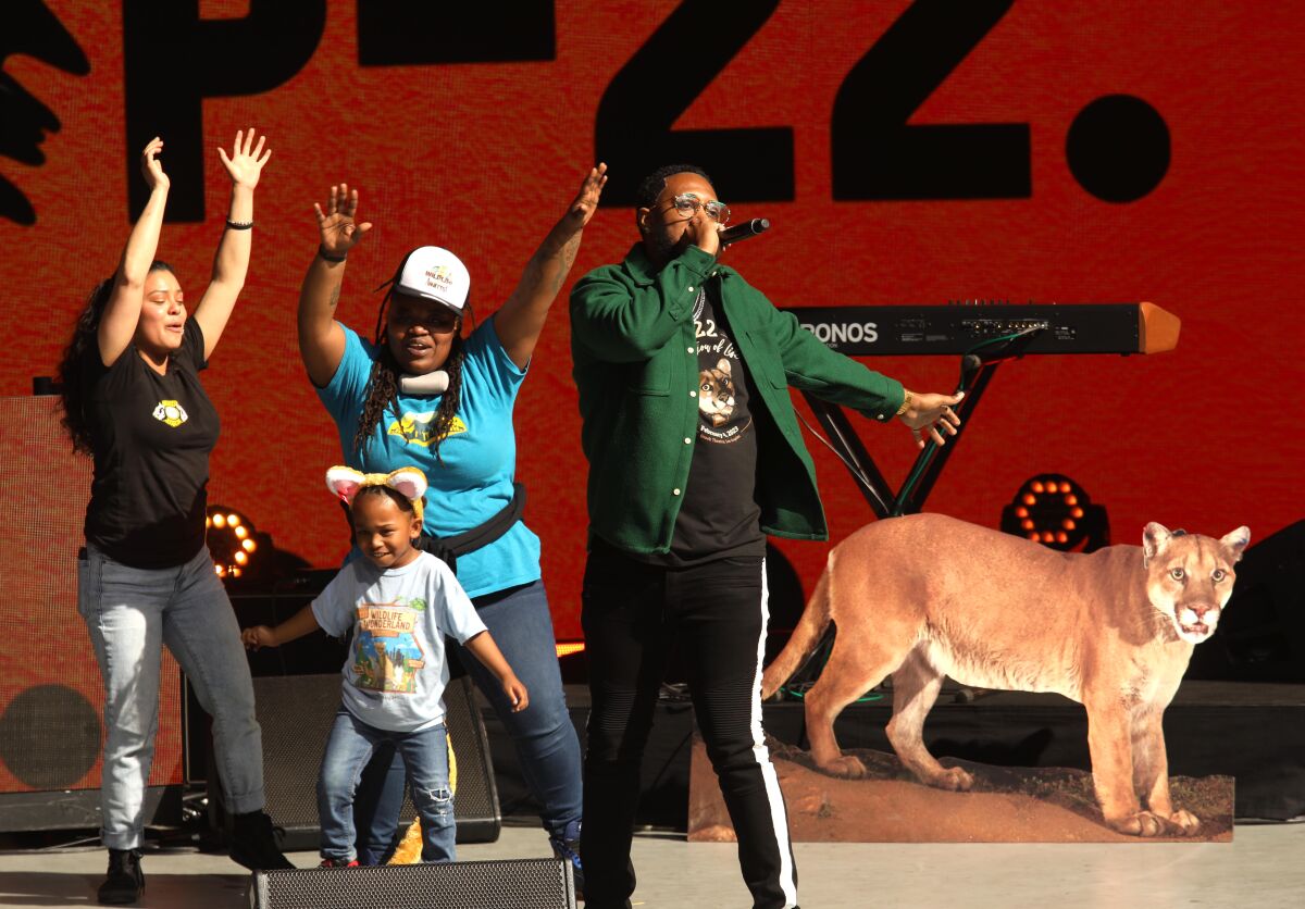 Two women, a man with a microphone, and a child wearing cat ears dance on stage with a cutout of a mountain lion behind them