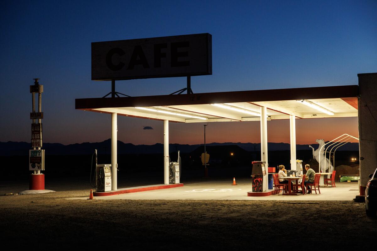 Two people sit under the awning of a gas station.