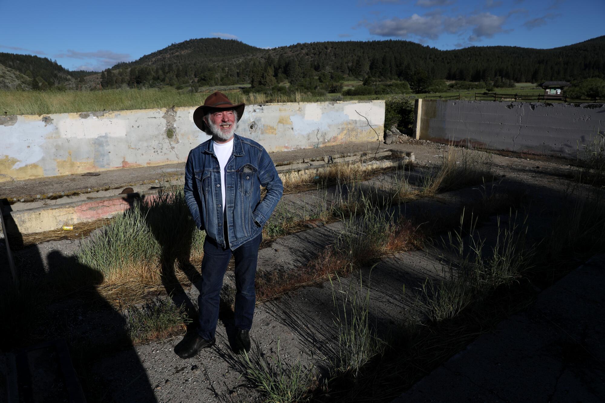 A man in a cowboy hat, beard and denim jacket stands in a grassy area surrounded by low walls.