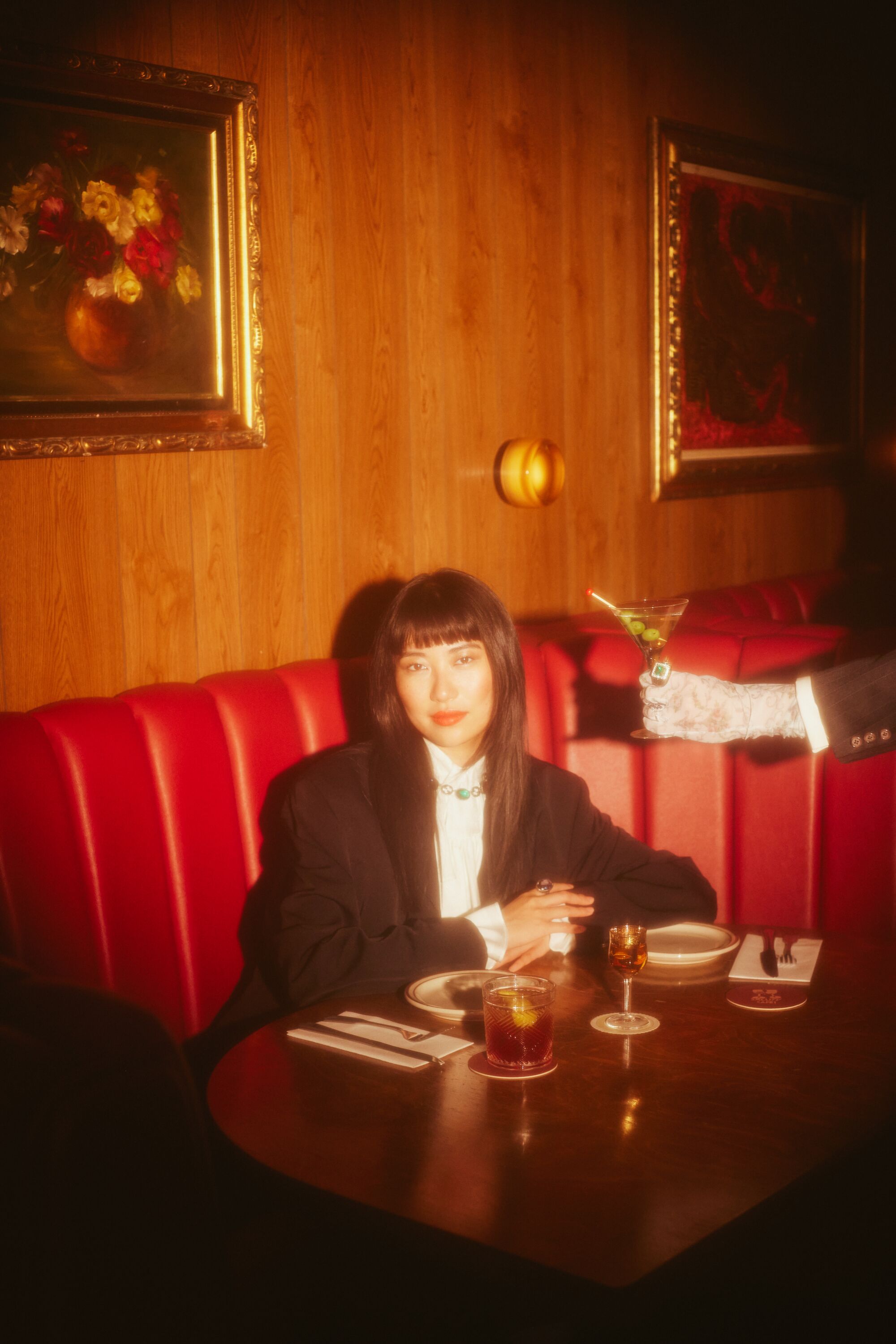 A gloved hand offers a martini to a woman sitting in a red booth.