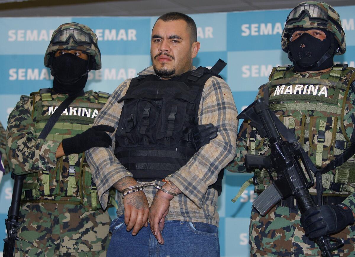 A handcuffed man wearing a ballistic vest is flanked by two Mexican troops in camouflage fatigues