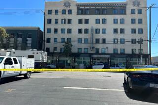 Police tape is seen in front of a parking lot. An older multi-story residential building is in the background.
