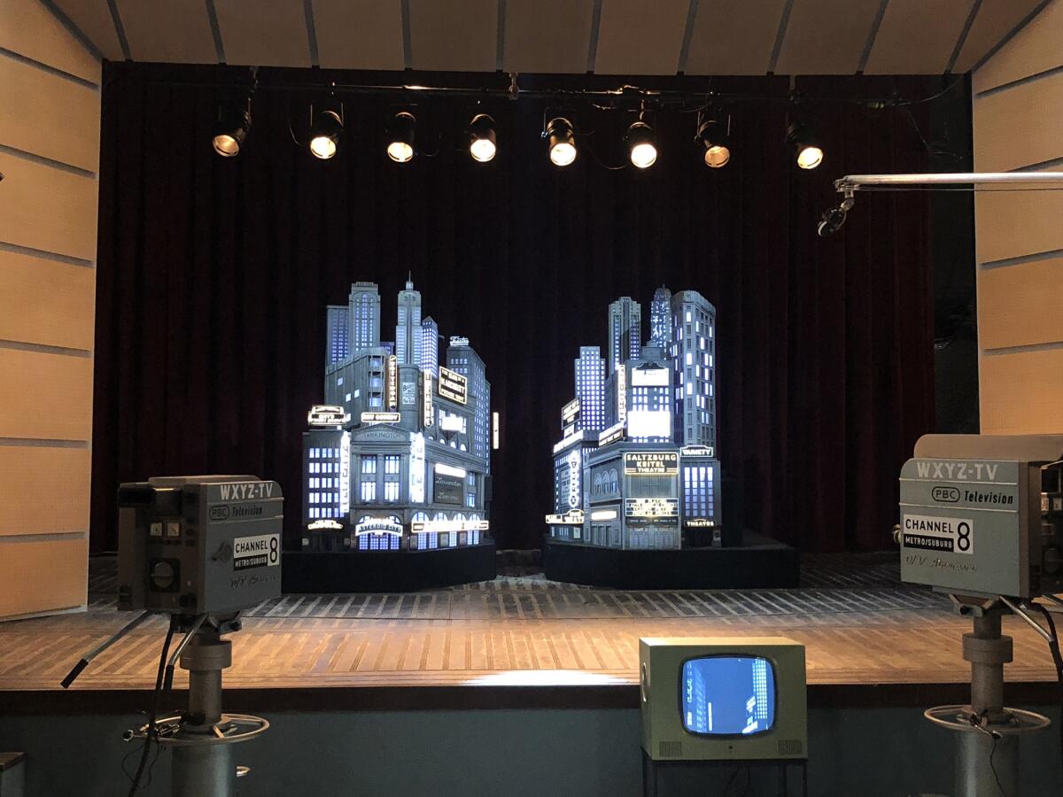 A stylized city skyline, nearly black and white (or gray), on a soundstage before old-timey TV cameras. 