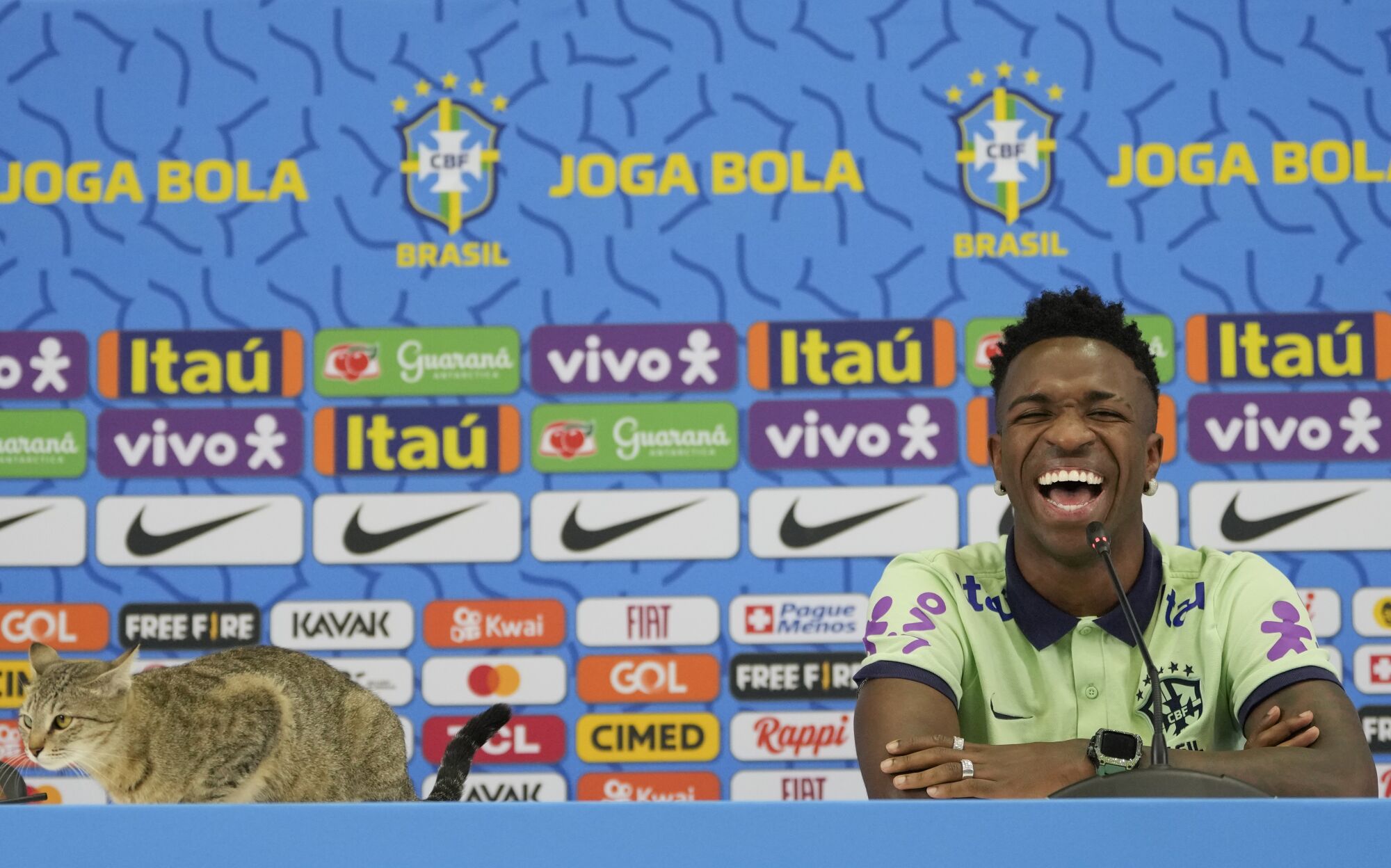 Brezilyalı Vinicius Junior, bir basın toplantısı sırasında masanın üzerine oturan bir kediye gülüyor.