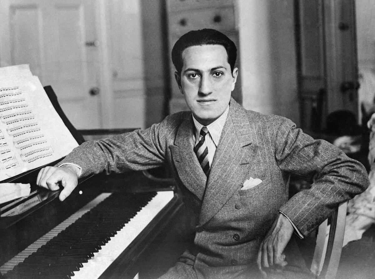 George Gershwin is seen sitting jauntily at a piano in a pinstripe suit.
