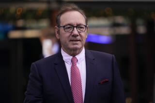 Kevin Spacey wearing glasses and in a dark suit, pink dress shirt and red tie standing against a blurred background