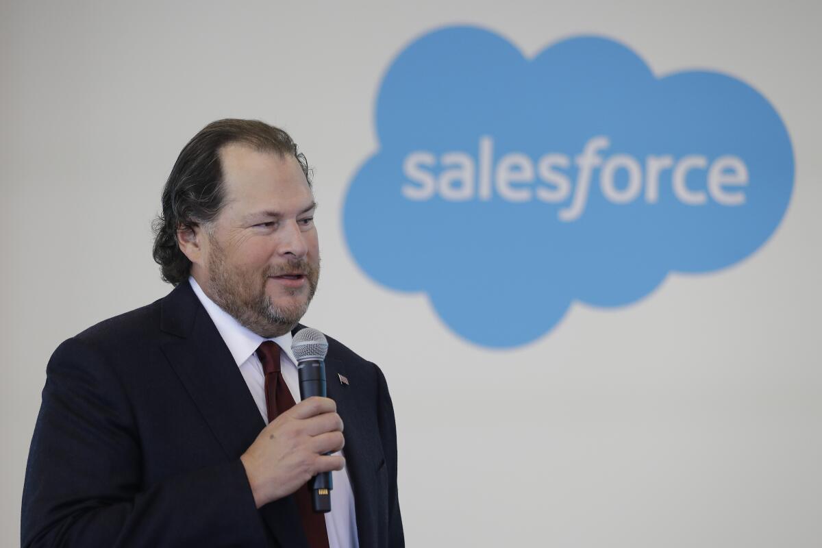 A man in a jacket and tie speaks using a microphone. To the right is a sign shaped like a blue cloud that says "salesforce." 