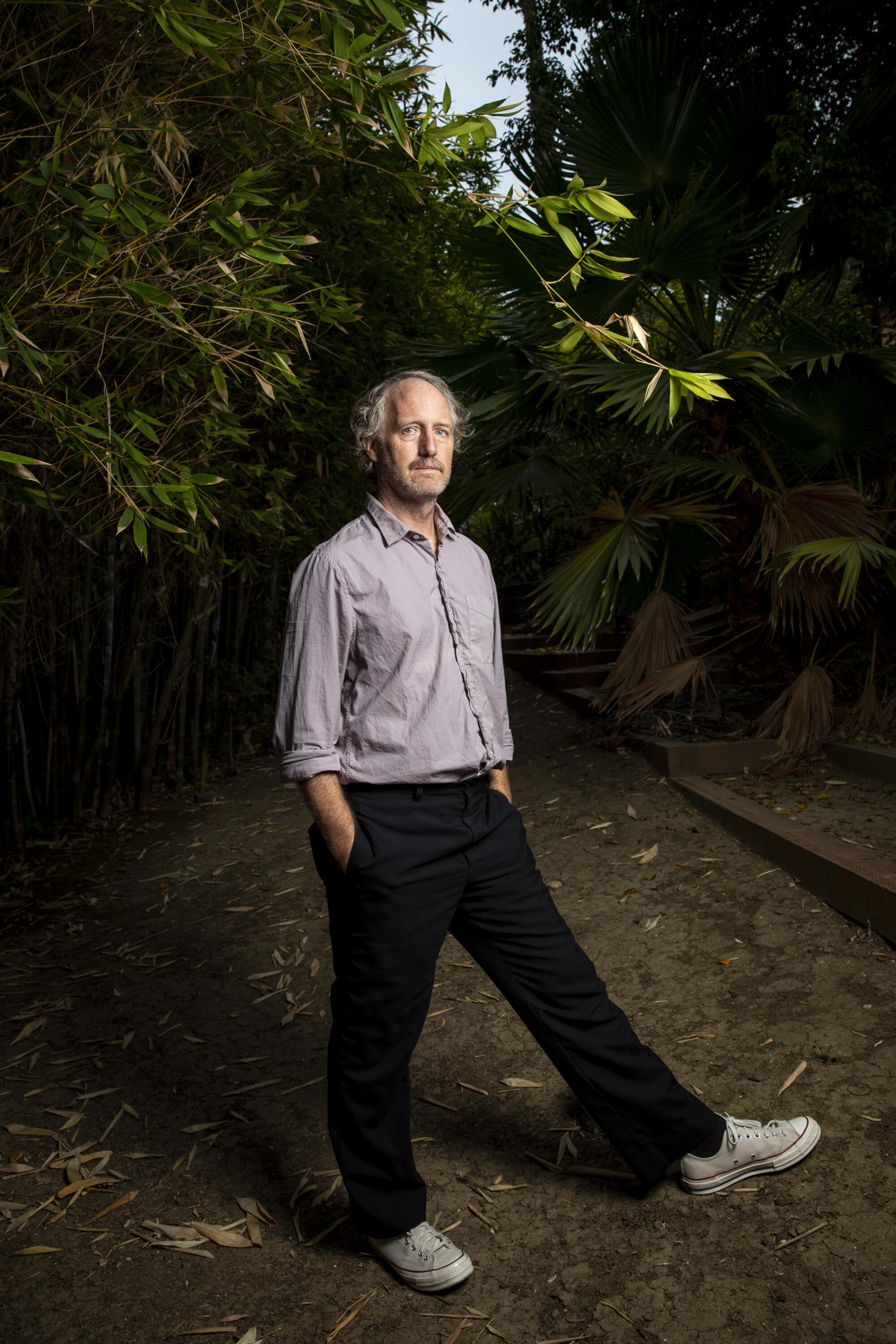 Writer and director Mike Mills stands outdoors on one foot, kicking up one leg.
