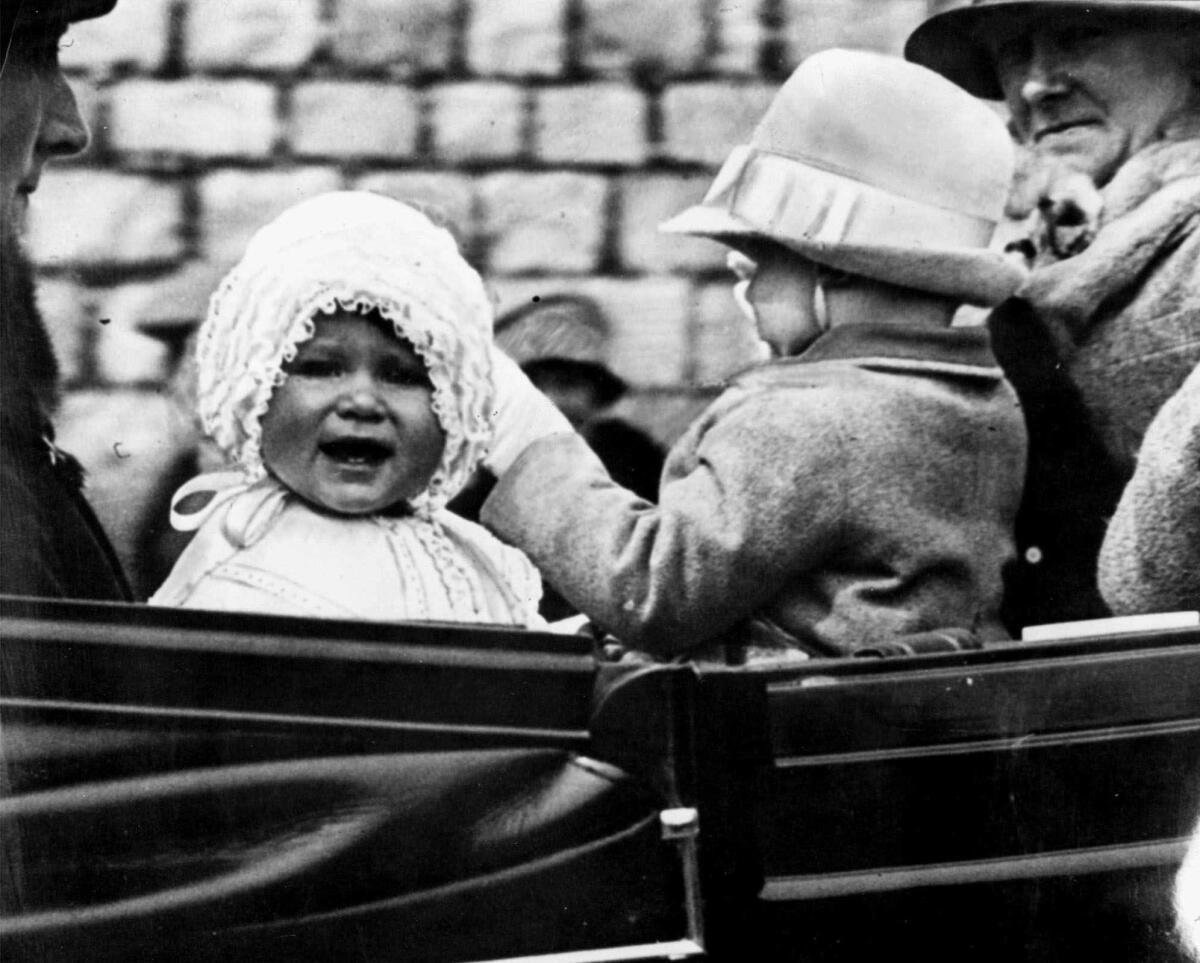 A baby in a frilly white bonnet sits in a vehicle with another child and an adult.