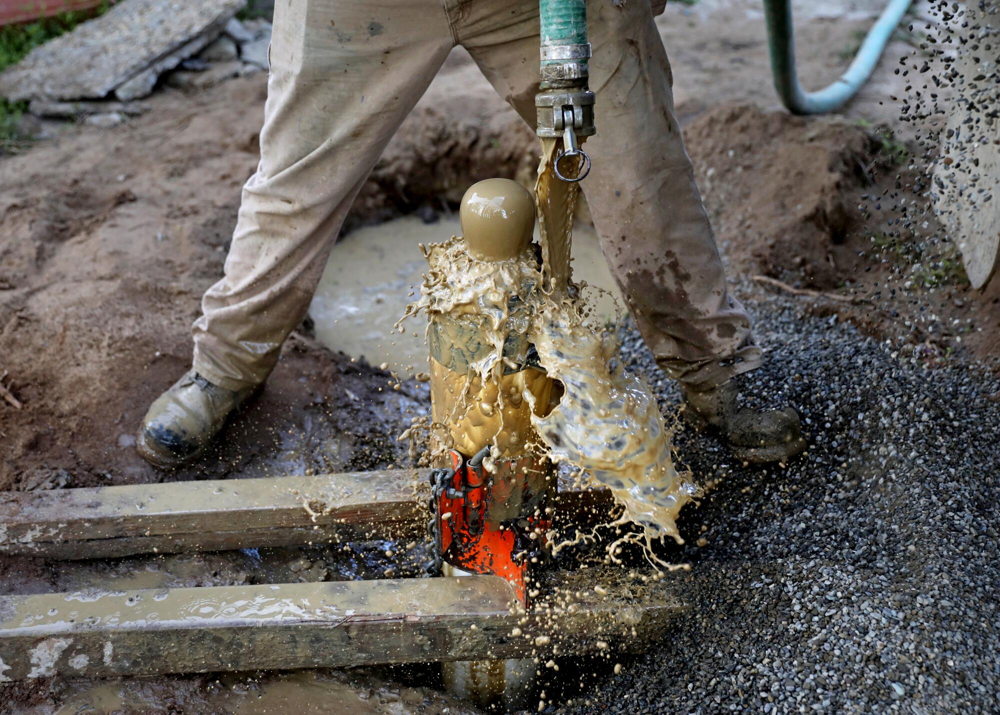 Dirty water splashes out of a hose as a person seen from the waist down drills a well. 